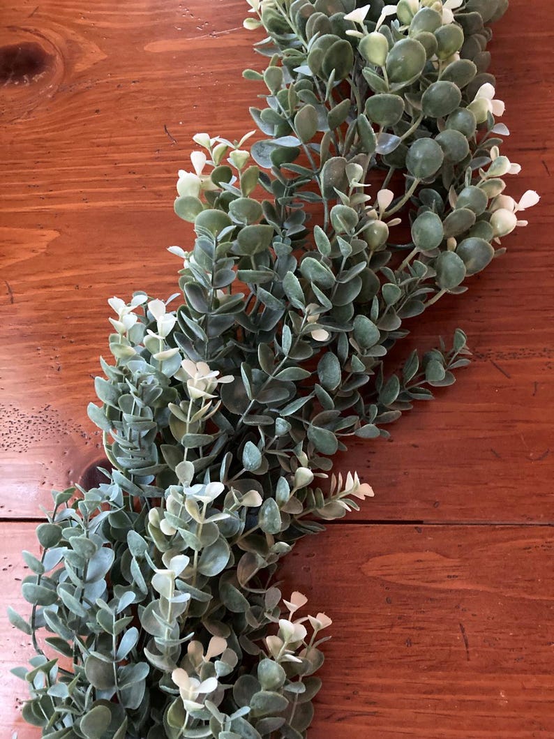 May include: A faux eucalyptus garland with green and white leaves. The garland is arranged on a wooden surface, creating a natural and decorative element. The leaves have a soft, textured appearance, adding a touch of greenery to any space.