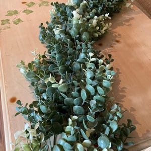 May include: A garland of artificial greenery with small, round, dark green leaves and lighter green accents. The garland is arranged in a flowing pattern, suitable for home decor or crafting projects. The leaves have a realistic appearance.