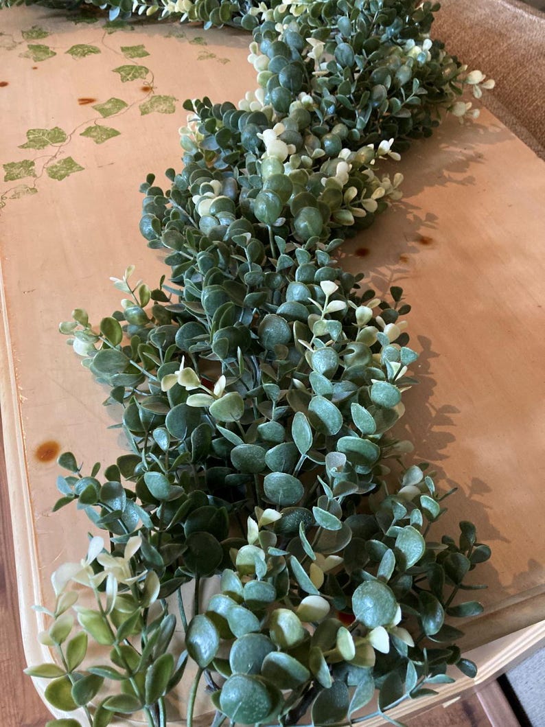 May include: A garland of artificial greenery with small, round, dark green leaves and lighter green accents. The garland is arranged in a flowing pattern, suitable for home decor or crafting projects. The leaves have a realistic appearance.