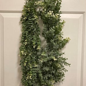 May include: A long, cascading garland of faux greenery, featuring small, round, green leaves with touches of white. The garland is hanging against a white door, creating a natural, decorative element for home decor.