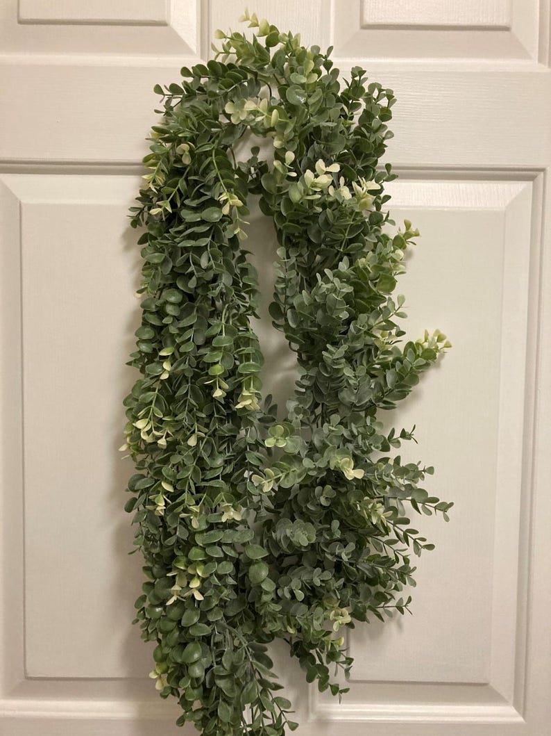May include: A long, cascading garland of faux greenery, featuring small, round, green leaves with touches of white. The garland is hanging against a white door, creating a natural, decorative element for home decor.
