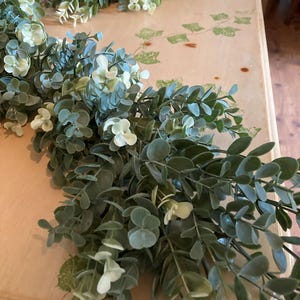 May include: Artificial eucalyptus garland with green and white leaves. The garland is arranged on a light-colored surface, creating a natural and decorative element. The leaves have a realistic appearance, suitable for home decor.