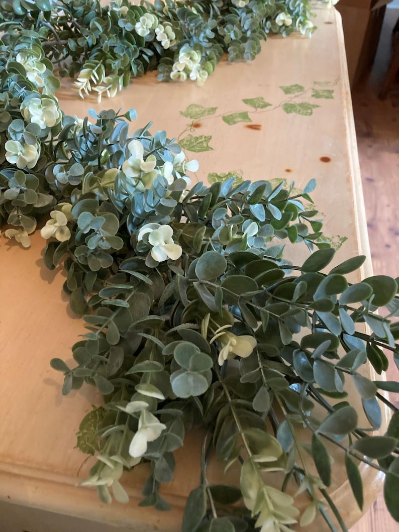 May include: Artificial eucalyptus garland with green and white leaves. The garland is arranged on a light-colored surface, creating a natural and decorative element. The leaves have a realistic appearance, suitable for home decor.