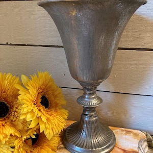 May include: A silver-colored metal urn with a flared top and a pedestal base. The urn is next to a bouquet of bright yellow sunflowers. The background is a light-colored wooden wall.