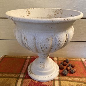 May include: A white pedestal planter with a wide, flared rim and a fluted, rounded body. The surface features a distressed, textured finish with brown speckles throughout, supported by a circular base with a beaded edge detail.