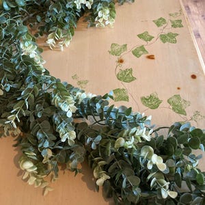 May include: A green and white eucalyptus garland, with small, round leaves, is draped across a light-colored surface. The garland is a mix of dark and light green, with some white accents. The surface has a vine pattern.