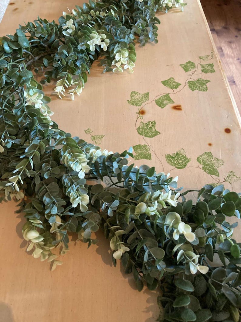 May include: A green and white eucalyptus garland, with small, round leaves, is draped across a light-colored surface. The garland is a mix of dark and light green, with some white accents. The surface has a vine pattern.