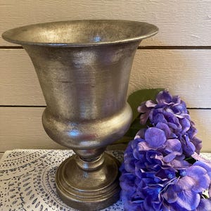 May include: A silver-toned urn with a wide, flared rim and a pedestal base. The urn has a bulbous midsection and a metallic finish. A cluster of vibrant purple hydrangea flowers rests beside the urn, adding a pop of color and texture.