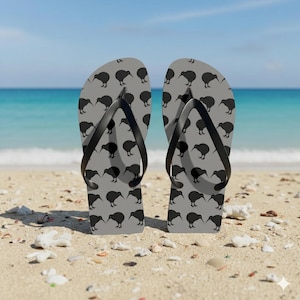 Może przedstawiać: Szare klapki z czarnym wzorem kiwi. Klapki mają czarne paski i leżą na piaszczystej plaży z morzem w tle. Modny i swobodny dodatek na lato.