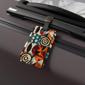 Pode incluir: Uma etiqueta de bagagem retangular com uma alça de couro preta e uma fivela de latão. A etiqueta apresenta um design de patchwork com formas geométricas em vermelho, laranja, azul e preto. A etiqueta está presa a uma mala cinza.