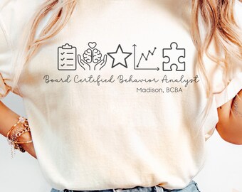 May include: Ivory-colored t-shirt with the text "Board Certified Behavior Analyst" and a series of black line art icons. The icons include a clipboard, brain, star, graph, and puzzle piece. The name "Madison, BCBA" is also printed on the shirt.