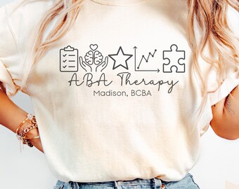 May include: Ivory-colored t-shirt with the text "ABA Therapy" and a series of black line-art icons. The icons include a clipboard, brain, star, graph, and puzzle piece. The text "Madison, BCBA" is below the main text.