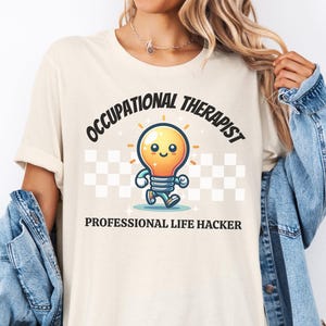 Może przedstawiać: Kremowa koszulka z napisem "OCCUPATIONAL THERAPIST" nad postacią z kreskówki w kształcie żarówki i napisem "PROFESSIONAL LIFE HACKER" poniżej. Żarówka jest pomarańczowa i żółta z niebieskimi nogami i ramionami. Tło ma wzór w szachownicę.
