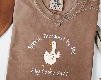 May include: A brown t-shirt with the text "Speech Therapist by day Silly Goose 24/7" and a cartoon goose illustration. The shirt is made of a soft, comfortable material.
