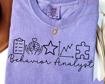 May include: A violet Comfort Colors t-shirt with the words "Behavior Analyst" and related graphics printed in black. The graphics include a clipboard, brain, star, graph, and puzzle piece. A gold necklace and a brown hat are also visible.