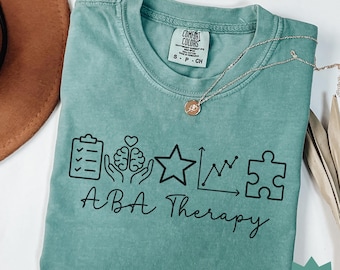May include: Seafoam green t-shirt with the words "ABA Therapy" and related graphic icons. A gold necklace with a pendant is draped across the shirt. The shirt is folded and has a tag that says "Comfort Colors".