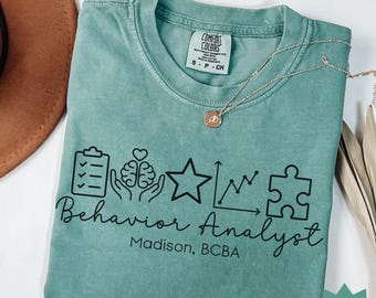 May include: Seafoam green t-shirt with the words "Behavior Analyst" and related graphic icons. The shirt also includes the text "Madison, BCBA". A silver necklace with a pendant is draped across the shirt.