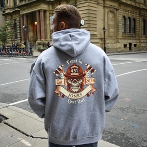 Puede incluir: Sudadera con capucha gris con un estampado gr&aacute;fico de un casco de bombero con una calavera y huesos cruzados. El texto "First in Last Out" est&aacute; impreso debajo del gr&aacute;fico. El texto "Est. 2025" est&aacute; impreso encima del gr&aacute;fico. El texto "Jones 451" est&aacute; impreso en el centro del gr&aacute;fico.