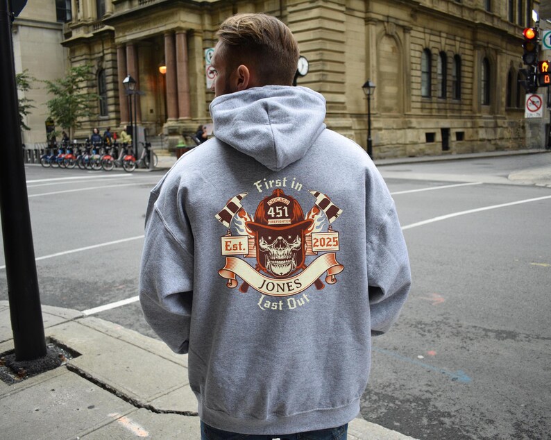 Puede incluir: Sudadera con capucha gris con un estampado gr&aacute;fico de un casco de bombero con una calavera y huesos cruzados. El texto "First in Last Out" est&aacute; impreso debajo del gr&aacute;fico. El texto "Est. 2025" est&aacute; impreso encima del gr&aacute;fico. El texto "Jones 451" est&aacute; impreso en el centro del gr&aacute;fico.