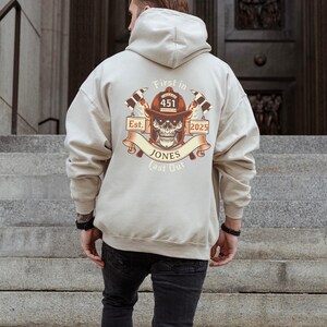 Puede incluir: Una sudadera con capucha beige con un gr&aacute;fico de una calavera con un casco de bombero con el texto "First In Last Out" y "Est. 2025" y el n&uacute;mero "451" en el casco.