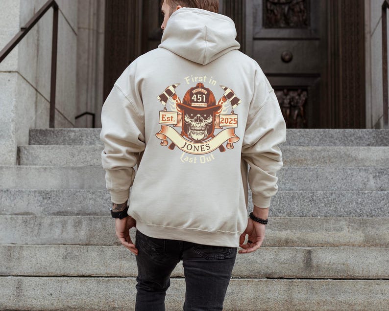 Puede incluir: Una sudadera con capucha beige con un gr&aacute;fico de una calavera con un casco de bombero con el texto "First In Last Out" y "Est. 2025" y el n&uacute;mero "451" en el casco.