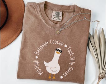 May include: A brown t-shirt with a white graphic of a goose wearing sunglasses and the text "Mo: High Behavior: CooCoo Tact: Silly Goose".