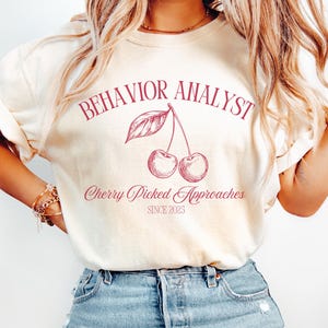 Könnte beinhalten: Cremefarbenes T-Shirt mit dem Schriftzug "BEHAVIOR ANALYST" in einem Bogen, und "Cherry Picked Approaches" unter einer Kirschen-Illustration. Der Text "SINCE 2023" steht unten. Das Shirt wird mit einer blauen Jeans getragen.
