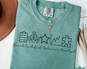May include: Seafoam green t-shirt with the text "Board Certified Behavior Analyst" and related graphic icons. The shirt has a crew neck and short sleeves. A silver necklace with a pendant is draped across the shirt.