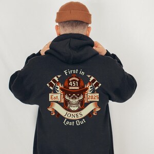 Puede incluir: Sudadera negra con un gr&aacute;fico de un casco de bombero con una calavera y huesos cruzados. El gr&aacute;fico incluye el texto "First in Last Out", "Chicago Firefighter", "Est. 2025", y "Jones".