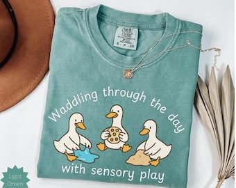 May include: Light green t-shirt with the text "Waddling through the day with sensory play" and three cartoon ducks. The ducks are interacting with sensory items. A silver necklace with a pendant is on the shirt.