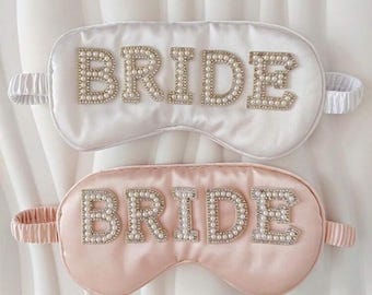 Op de afbeelding: Twee satijnen slaapmaskers, één witte en één blush roze, elk geborduurd met het woord "BRIDE" in parelachtige kralen. De maskers hebben elastische bandjes voor comfortabel dragen en zijn ontworpen voor bruidsfeesten.