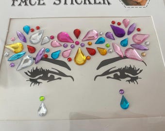 Op de afbeelding: Een vel gezichtsstickers met een ontwerp van gestileerde ogen en kleurrijke strass-steentjes. De strass-steentjes zijn in verschillende vormen en kleuren, waaronder roze, blauw, geel en paars. Het stickervel is wit.