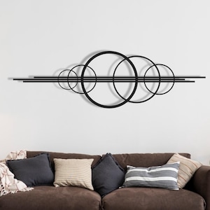 Puede incluir: Arte mural de metal negro con círculos superpuestos y líneas rectas. El diseño moderno está montado sobre un sofá marrón con cojines y una manta. Una lámpara de pie y una planta en maceta completan la decoración de la habitación.