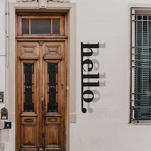 Op de afbeelding: Een zwart metalen bord met het woord "hello" verticaal op een witte muur. Het bord staat naast een houten deur en een raam met groene luiken. De deur is van donker hout en heeft ingewikkelde details.