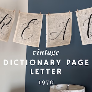 May include: A banner spelling "READ" made from vintage dictionary pages, with black letters. The pages are suspended from a string with small wooden pegs. The background is a two-tone wall, with white and navy blue paint. The text "vintage DICTIONARY PAGE LETTER 1970" is also visible.