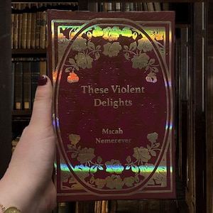 Puede incluir: Un libro burdeos titulado "These Violent Delights" de Micah Nemerever. La portada presenta un dise&ntilde;o ovalado con rosas doradas y un borde hologr&aacute;fico. El fondo muestra una biblioteca con estanter&iacute;as.
