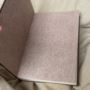 Puede incluir: Diario abierto con una cubierta marr&oacute;n y p&aacute;ginas con estampado floral. Las p&aacute;ginas son de color beige claro con un dise&ntilde;o floral delicado y repetitivo. El diario est&aacute; abierto, revelando las p&aacute;ginas interiores.