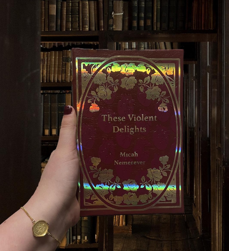 Puede incluir: Un libro burdeos titulado "These Violent Delights" de Micah Nemerever. La portada presenta un dise&ntilde;o ovalado con rosas doradas y un borde hologr&aacute;fico. El fondo muestra una biblioteca con estanter&iacute;as.