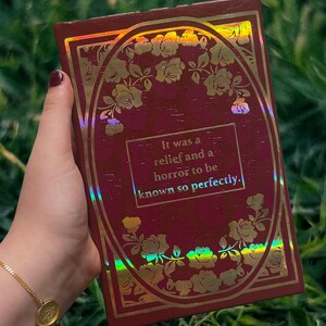 Puede incluir: Un libro de color burdeos con detalles florales dorados y la cita: "It was a relief and a horror to be known so perfectly." La portada tiene un marco ovalado dorado con rosas y un efecto hologr&aacute;fico.