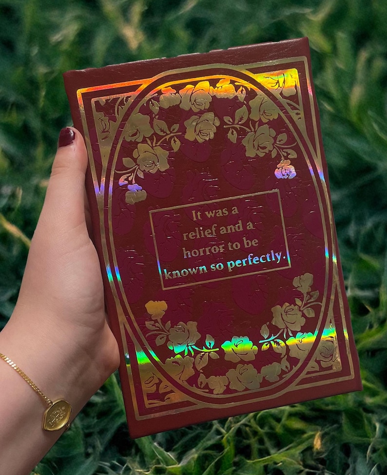 Puede incluir: Un libro de color burdeos con detalles florales dorados y la cita: "It was a relief and a horror to be known so perfectly." La portada tiene un marco ovalado dorado con rosas y un efecto hologr&aacute;fico.