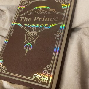 Puede incluir: Un libro de tapa dura titulado "The Prince" con una cubierta marr&oacute;n y detalles dorados. El t&iacute;tulo est&aacute; en una fuente decorativa, y la cubierta tiene dise&ntilde;os de borde ornamentados. El libro est&aacute; sobre una superficie de tela beige.