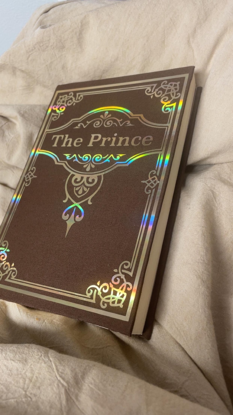 Puede incluir: Un libro de tapa dura titulado "The Prince" con una cubierta marr&oacute;n y detalles dorados. El t&iacute;tulo est&aacute; en una fuente decorativa, y la cubierta tiene dise&ntilde;os de borde ornamentados. El libro est&aacute; sobre una superficie de tela beige.