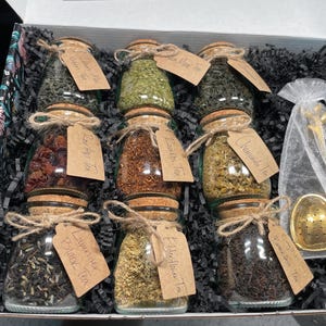 Könnte beinhalten: Geschenkbox mit Gläsern mit losem Blatttee, jeweils mit einem Korken verschlossen und mit Bindfaden gebunden. Die Gläser sind mit Teesorten wie Lavendel-Schwarztee und Kamillentee beschriftet. Ein goldener Tee-Infuser ist enthalten.