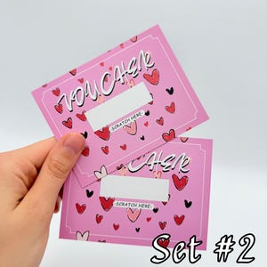 May include: Two pink scratch-off cards with a heart pattern and the text "Voucher" and "Check" with the words "Scratch Here" below each. The cards are labeled "Set #2".