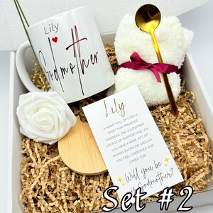 May include: A gift set in a white box with shredded paper includes a white mug with "Lily Godmother" in script, a white rose, a small white towel tied with a burgundy ribbon, a gold spoon, and a card that says "Will you be my Godmother?"