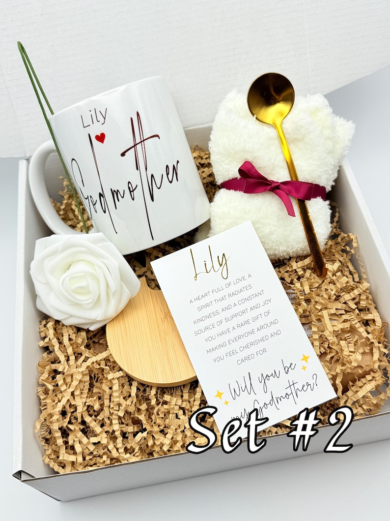 May include: A gift set in a white box with shredded paper includes a white mug with "Lily Godmother" in script, a white rose, a small white towel tied with a burgundy ribbon, a gold spoon, and a card that says "Will you be my Godmother?"