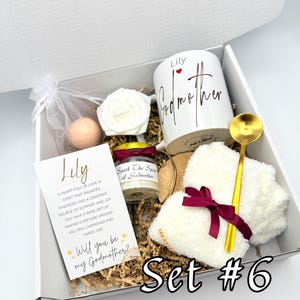 May include: A gift box with a white mug, a candle, a card, a rose, a bath bomb, a gold spoon, and a white fluffy item tied with a burgundy ribbon. The mug says "Lily Godmother". The card says "Will you be my Godmother?" and "Set #6".