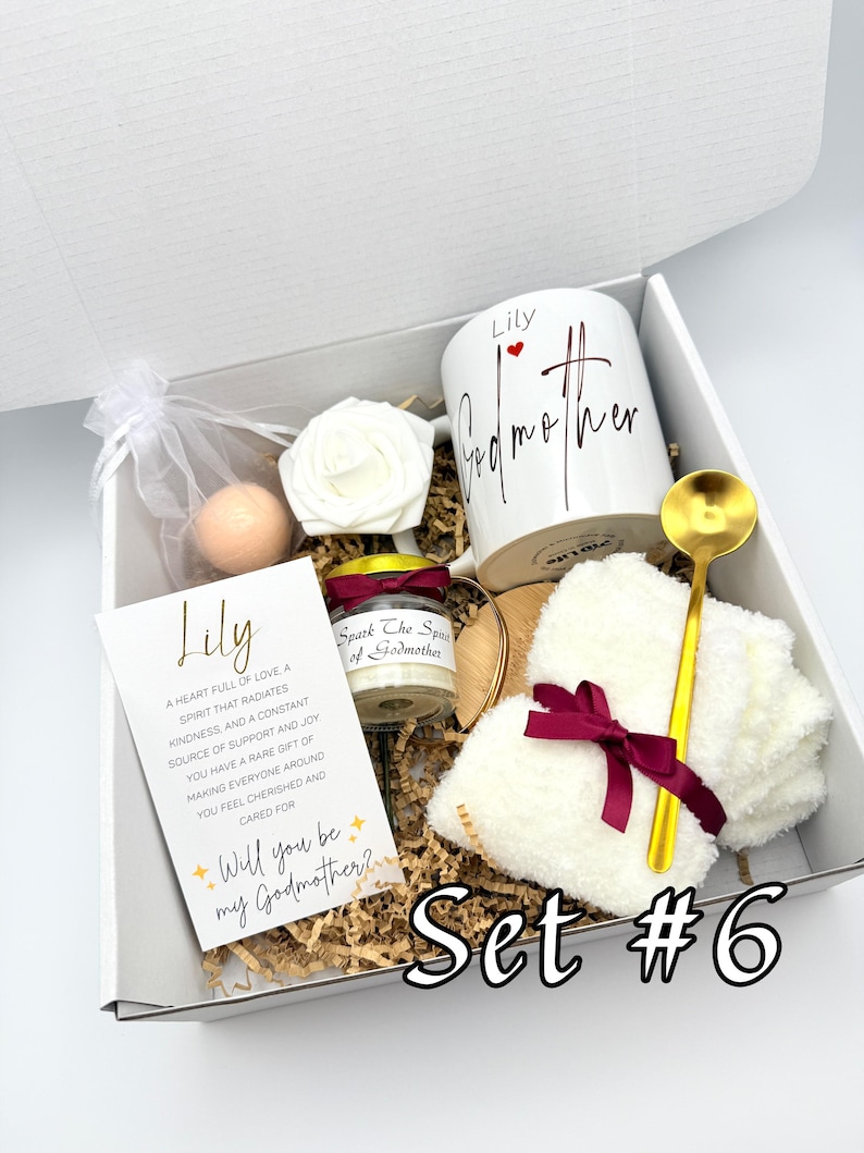 May include: A gift box with a white mug, a candle, a card, a rose, a bath bomb, a gold spoon, and a white fluffy item tied with a burgundy ribbon. The mug says "Lily Godmother". The card says "Will you be my Godmother?" and "Set #6".