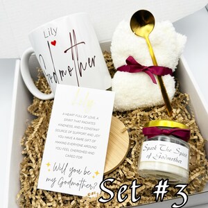 May include: A gift set in a white box with shredded paper includes a white mug with "Lily Godmother" text, a small candle, a gold spoon, and a white washcloth tied with a burgundy ribbon. A card reads "Will you be my Godmother?"