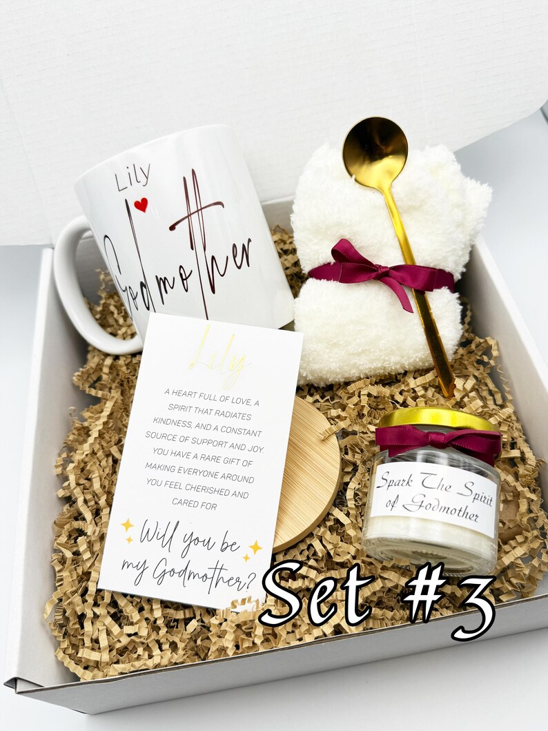 May include: A gift set in a white box with shredded paper includes a white mug with "Lily Godmother" text, a small candle, a gold spoon, and a white washcloth tied with a burgundy ribbon. A card reads "Will you be my Godmother?"
